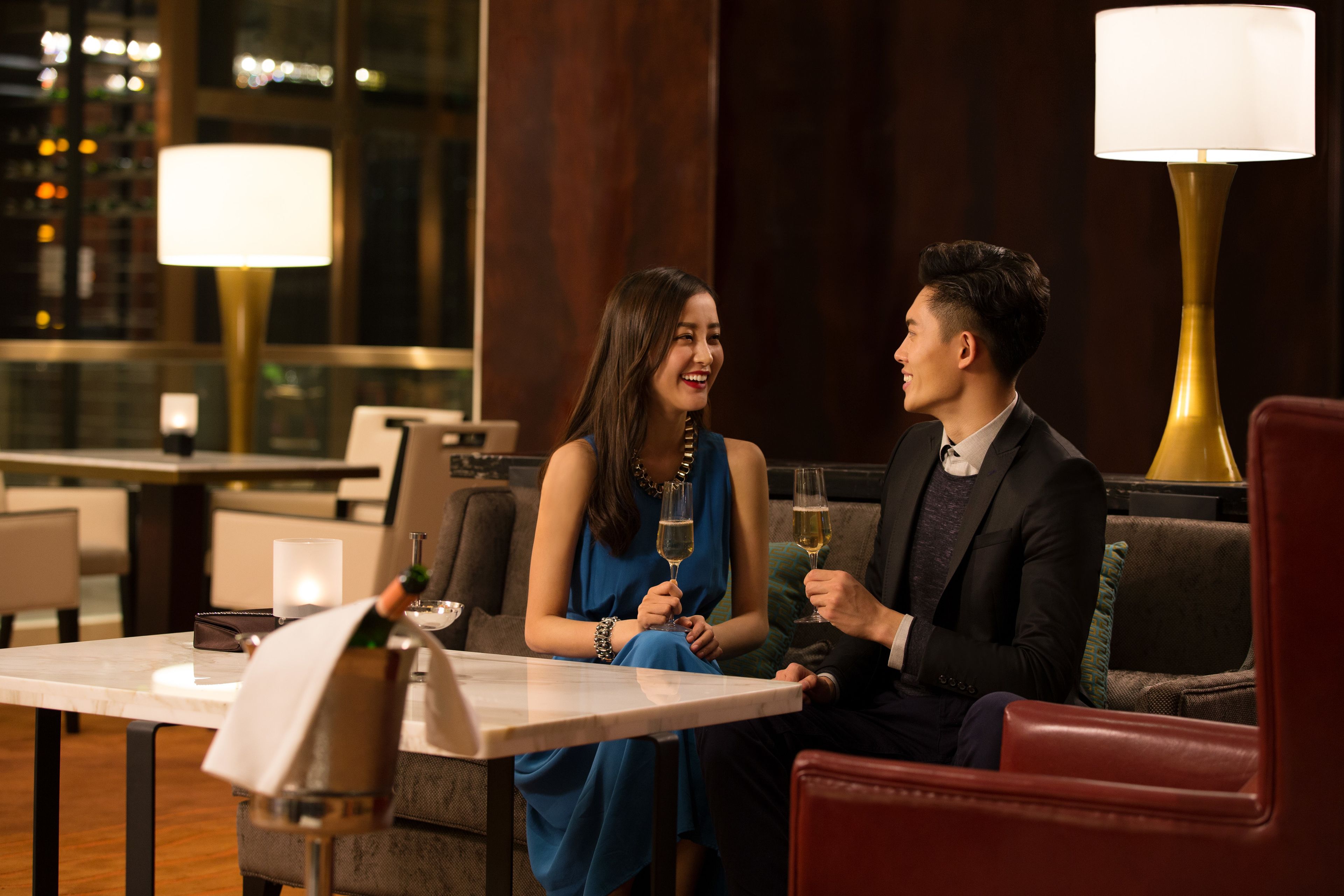 Una pareja sentada a una mesa en un ambiente interior bien iluminado, con vista a la ciudad de noche a través de amplias ventanas.