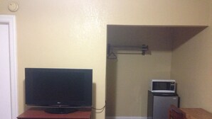 A room with a television on a wooden dresser, a microwave on a small table, and a shelf with a hanging object. - Ole English Inn (Tuscaloosa)