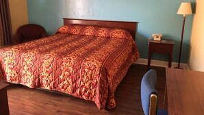 A hotel room with a wooden bed, a blue wall, a framed picture, a bedside table with a lamp, and a chair. - Ole English Inn (Tuscaloosa)