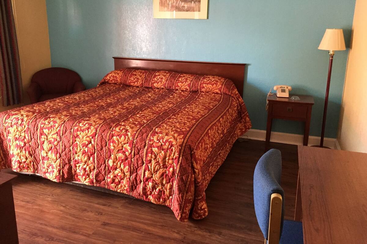 Een hotelkamer met een houten bed, een blauwe wand, een lijst met foto, een nachtkastje met een lamp en een stoel.