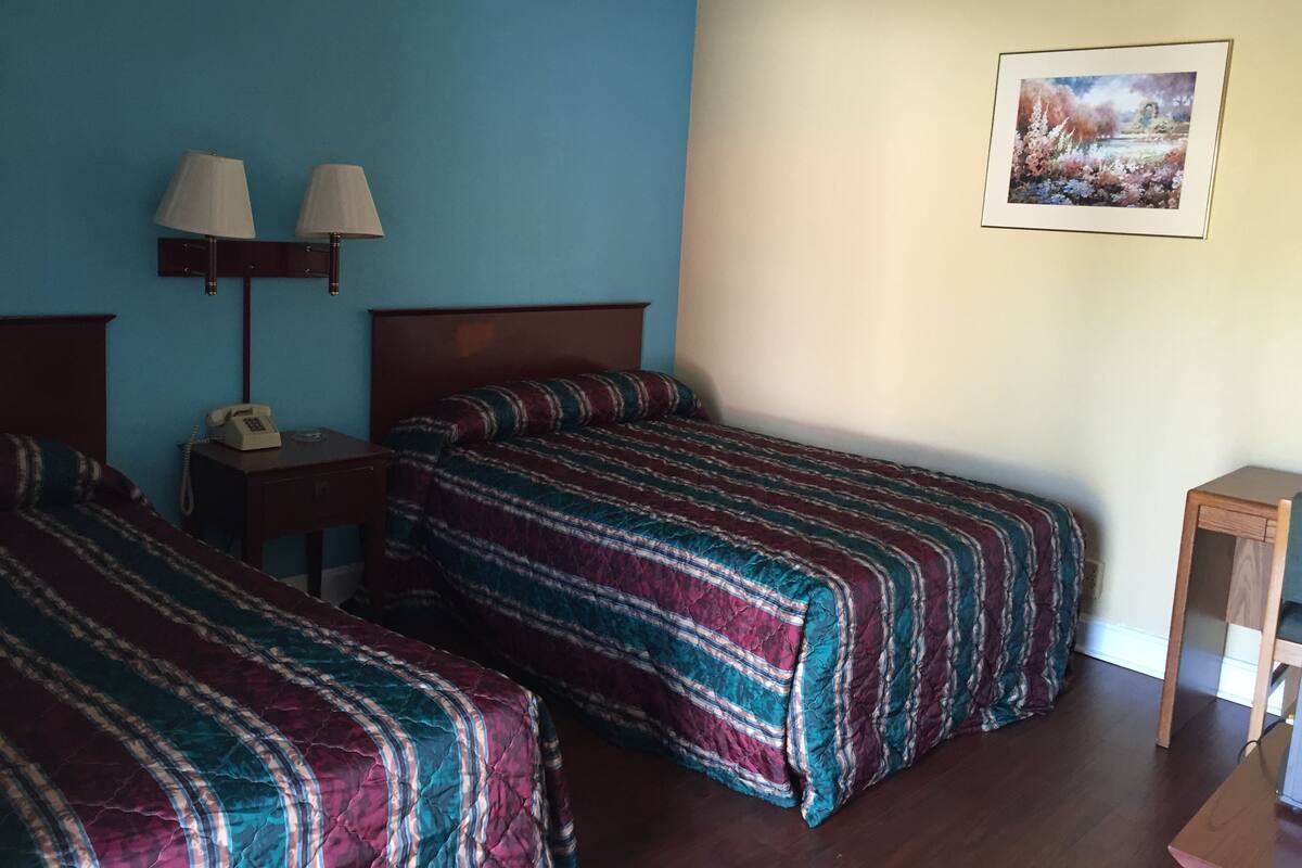 Een kamer met twee bedden, een nachtkastje met een lamp, een klein tafeltje en een ingelijst schilderij aan de muur.