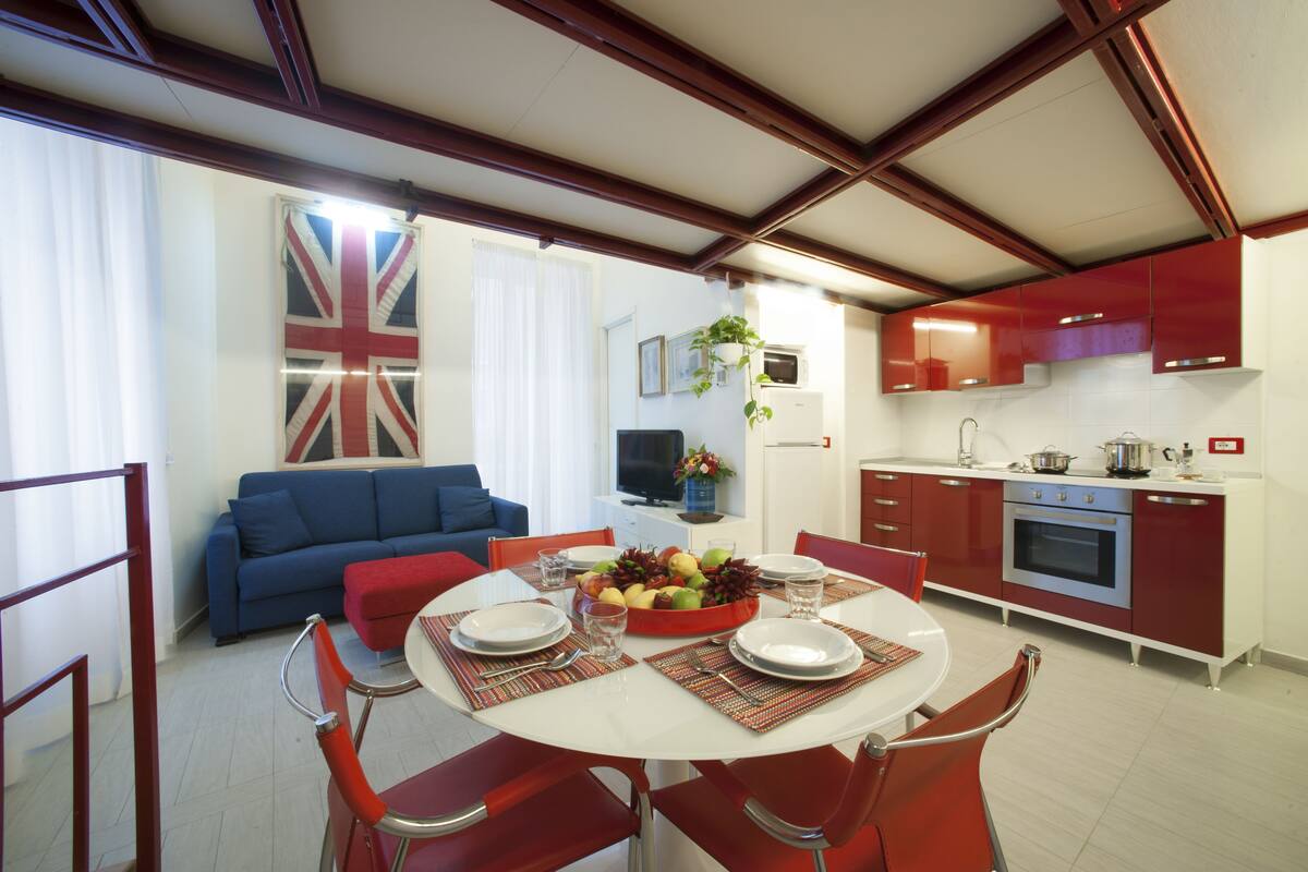Un coin repas avec une table ronde dressée pour quatre personnes, entourée de chaises rouges. Un canapé bleu et un fauteuil rouge sont visibles à l’arrière-plan.