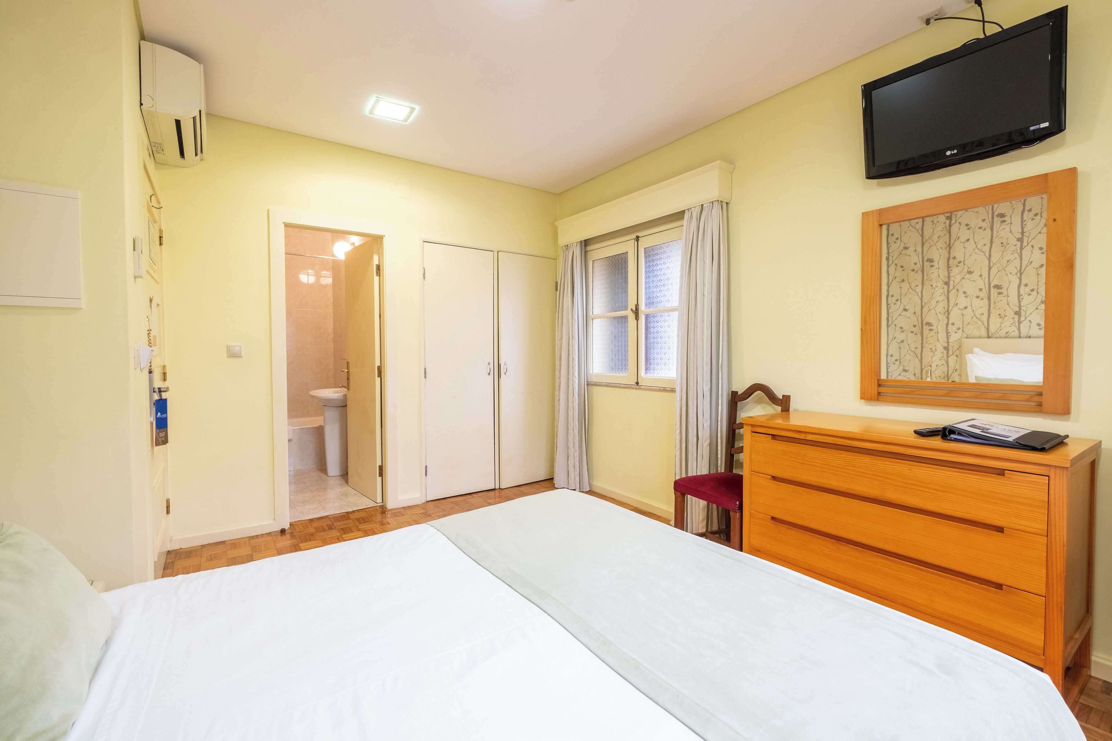 Ein Schlafzimmer mit Bett, einem hölzernen Schminktisch, einem Fernseher, einem Fenster mit Vorhängen und einer Tür zum Badezimmer.
