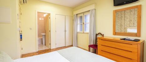 Ein Schlafzimmer mit Bett, einem hölzernen Schminktisch, einem Fernseher, einem Fenster mit Vorhängen und einer Tür zum Badezimmer.