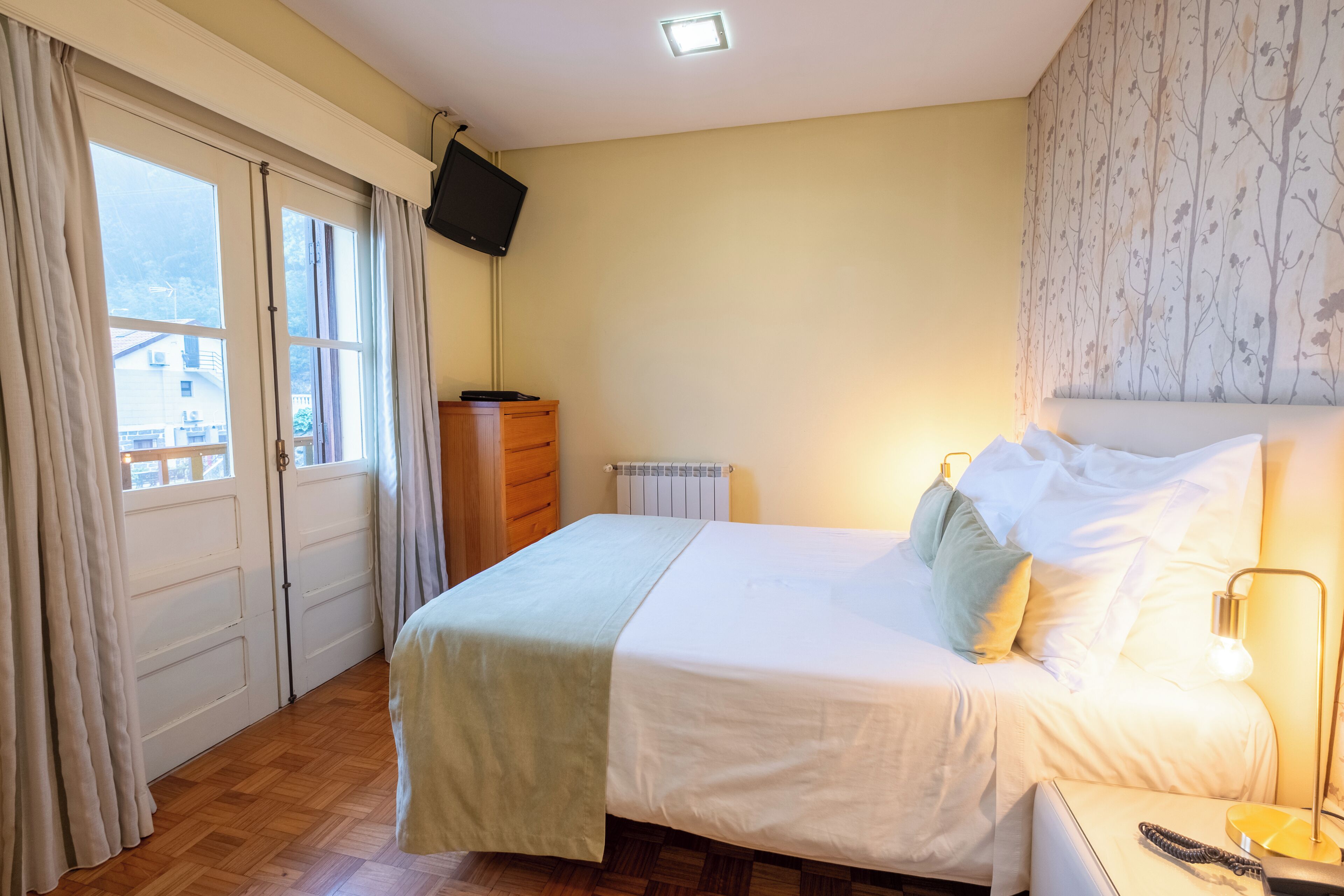 A bedroom with a bed, a dresser, a TV mounted on the wall, and a window with curtains.