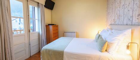 A bedroom with a bed, a dresser, a TV mounted on the wall, and a window with curtains.
