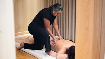 Iemand ontvangt een rugmassage op een mat in een kamer met houten vloeren en een raam met zonwering.