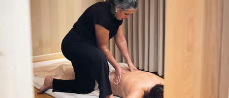 A person receiving a back massage on a mat in a room with wooden flooring and a window with blinds.