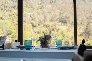A table set with blue glassware, a glass bowl with a plant, and a knife on a table with a white tablecloth, overlooking a forested landscape. - Adelaide Hotel (Terras de Bouro)