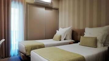 A hotel room with two beds, a wardrobe, an air conditioning unit, and a window with curtains.