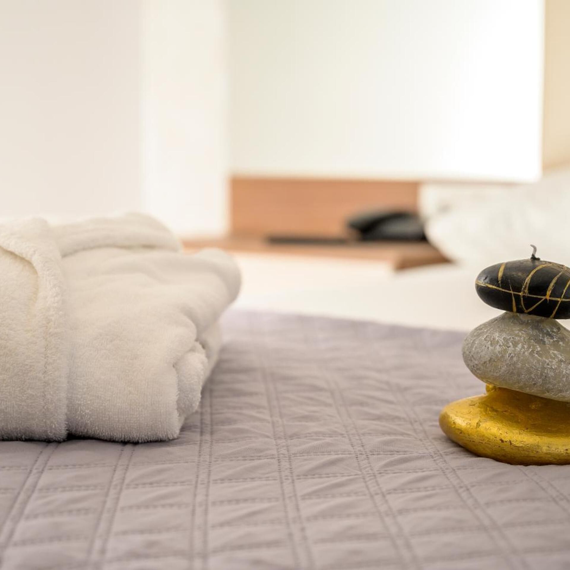 A neatly made bed with a white blanket, a grey quilt, and a stack of smooth stones on a golden base.
