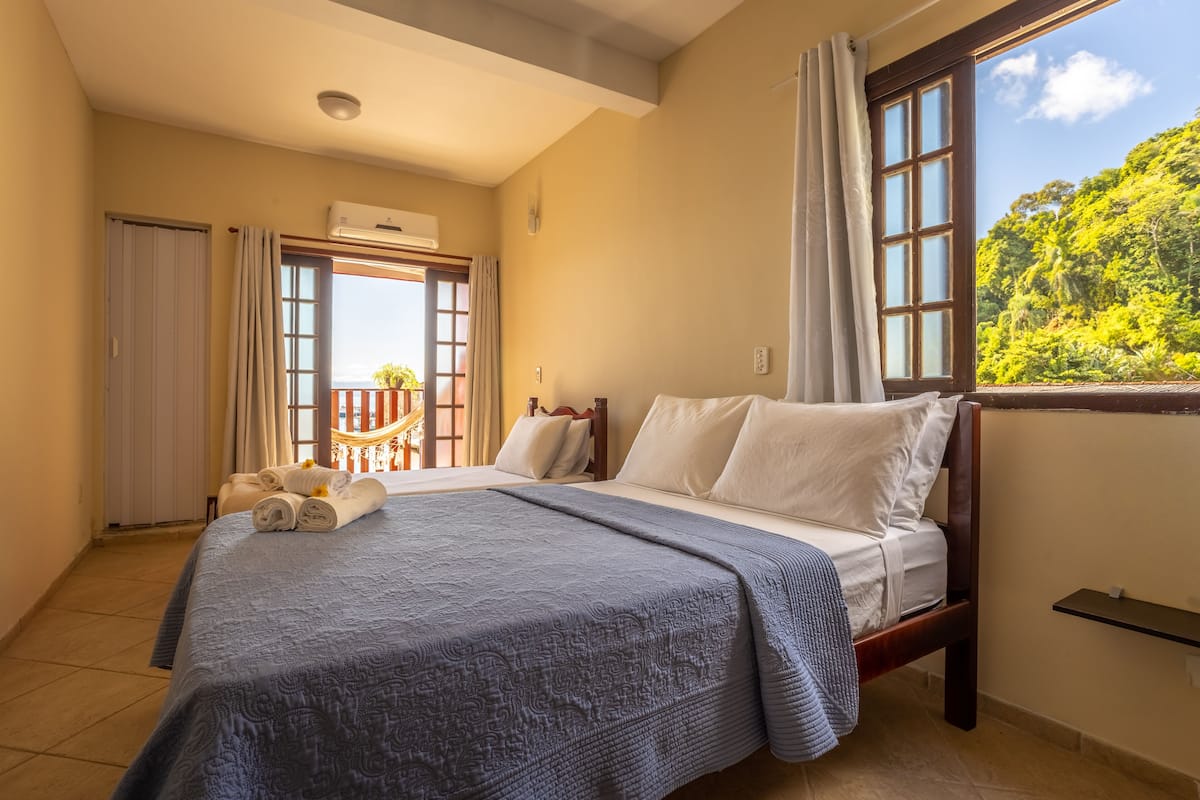 A hotel room with a large bed, a balcony with a view of greenery, and a window with curtains.