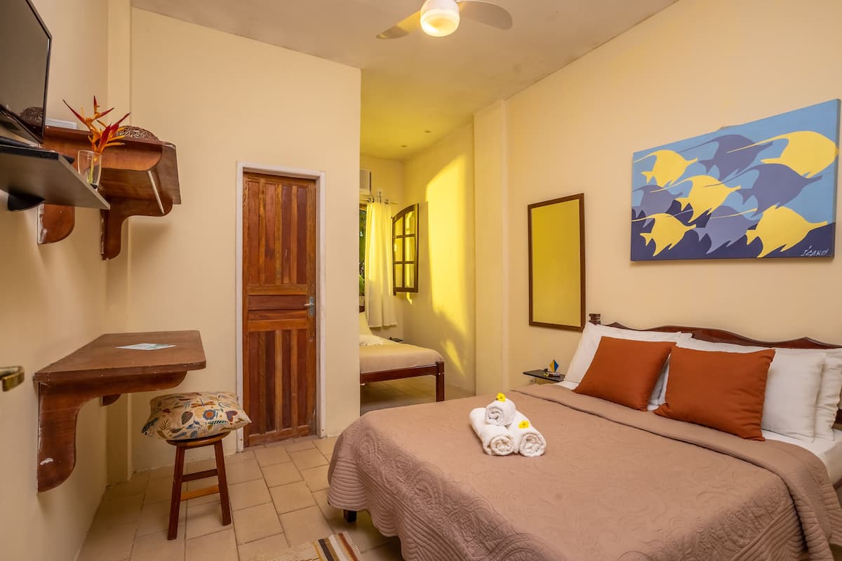 A hotel room with a bed, a desk, a chair, a wooden door, and a painting on the wall.