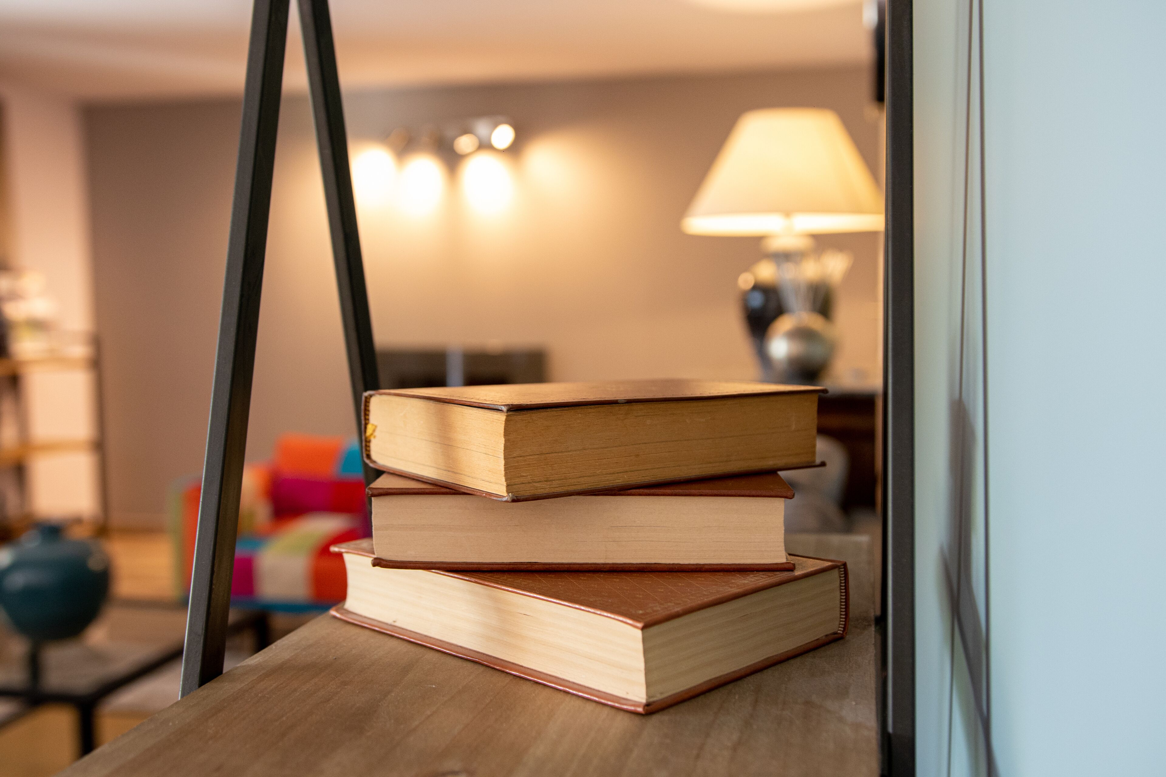A stack of books on a wooden table in a room with a lamp and a colorful chair in the background.