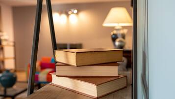 A stack of books on a wooden table in a room with a lamp and a colorful chair in the background.