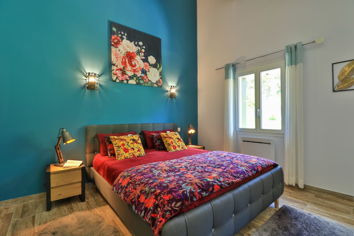 Un dormitorio con una cama grande, una mesita de noche, una lámpara de pared, una ventana con cortinas y un cuadro floral en la pared.