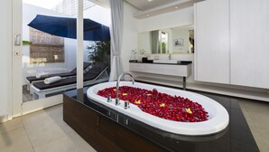 A modern bathroom with a large bathtub filled with red rose petals, a glass-enclosed seating area with a blue umbrella, and a large mirror above the sink. - Bajra Bali Villa (Seminyak)