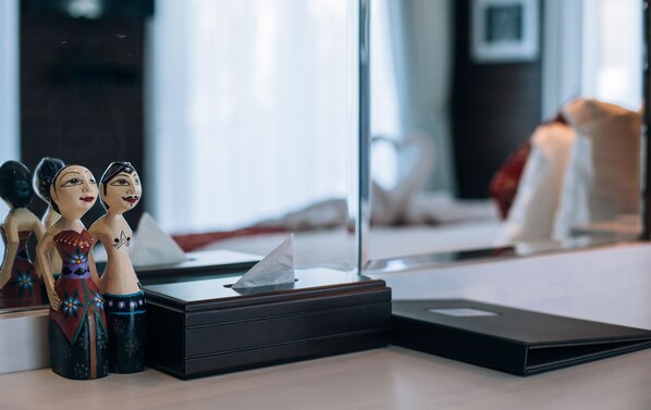 A room with a mirror reflecting a bed and a window. There are two wooden figurines on a table.