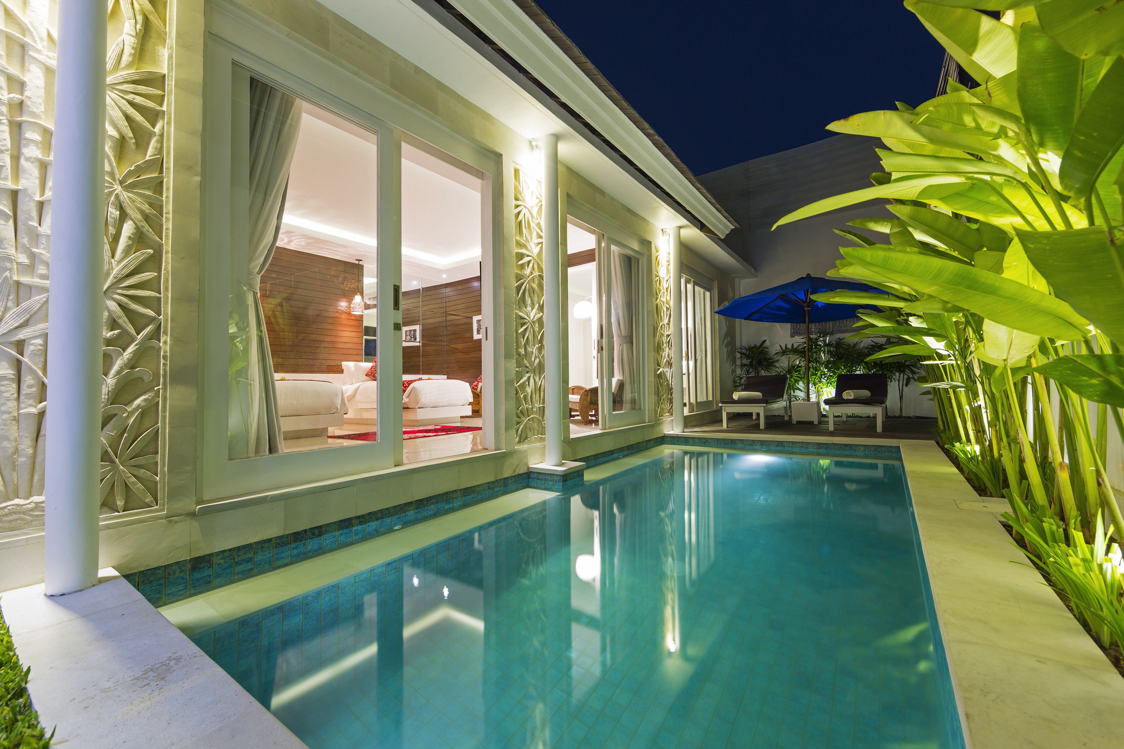 A swimming pool with a glass enclosure, a covered seating area, and a view of a bedroom with a red bedspread.