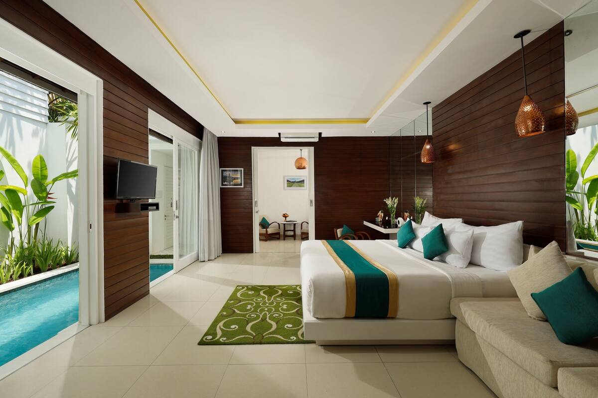 A modern hotel room with a large bed, a sofa, a small pool, and a dining area visible through an open door.