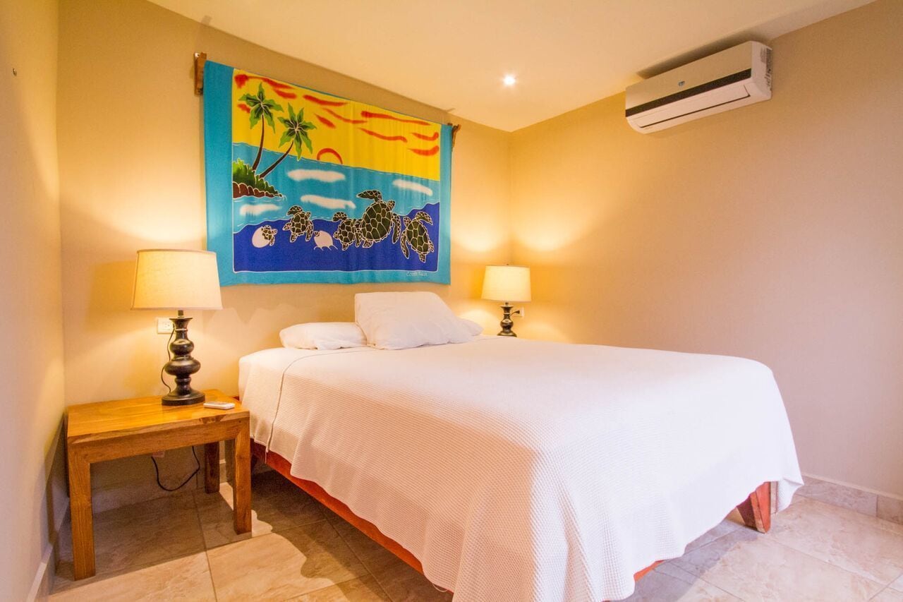 A bedroom with a bed, two bedside lamps, a wall tapestry, and an air conditioning unit.