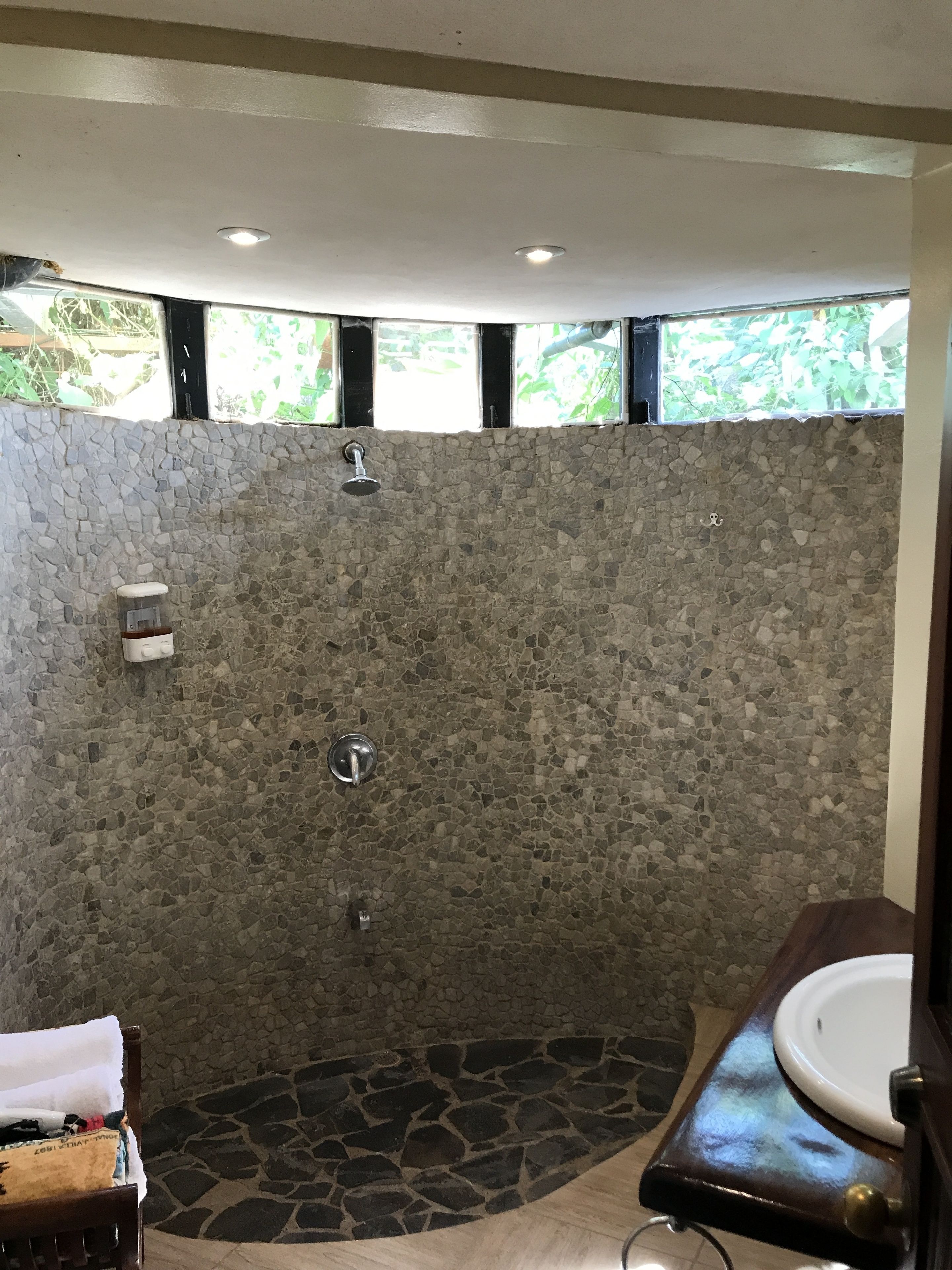 Une salle de bain avec un mur de douche en pierre, un lavabo et une chaise en bois.