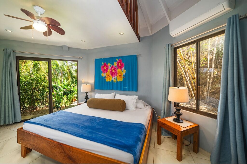 A bedroom with a bed, a ceiling fan, a window with curtains, and a wall with a floral painting.
