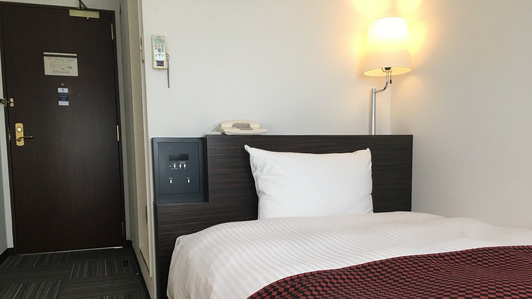 A hotel room with a bed, a bedside telephone, a wall-mounted lamp, and a door with a lock.