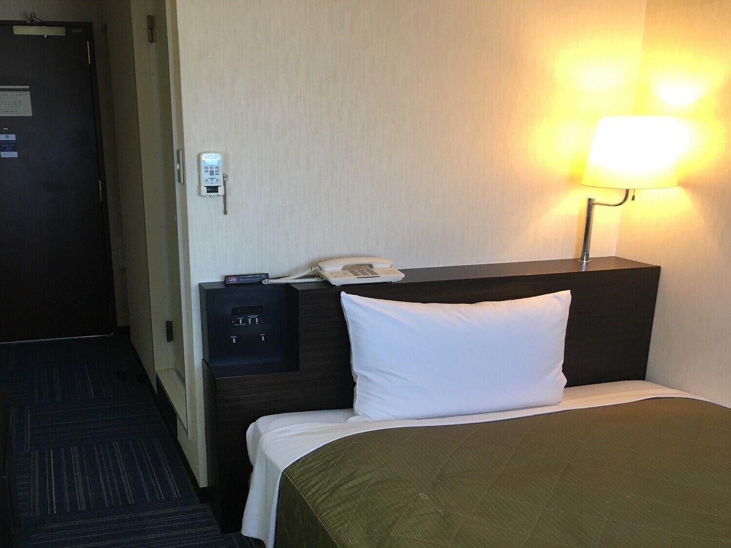 A hotel room with a bed, a bedside table with a lamp, a phone, and a control panel.