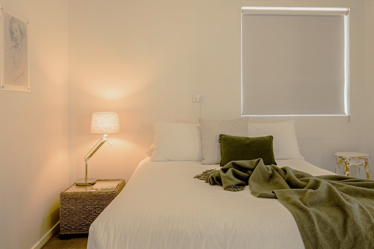 A bedroom with a bed, a bedside lamp, a window, and a small stool.