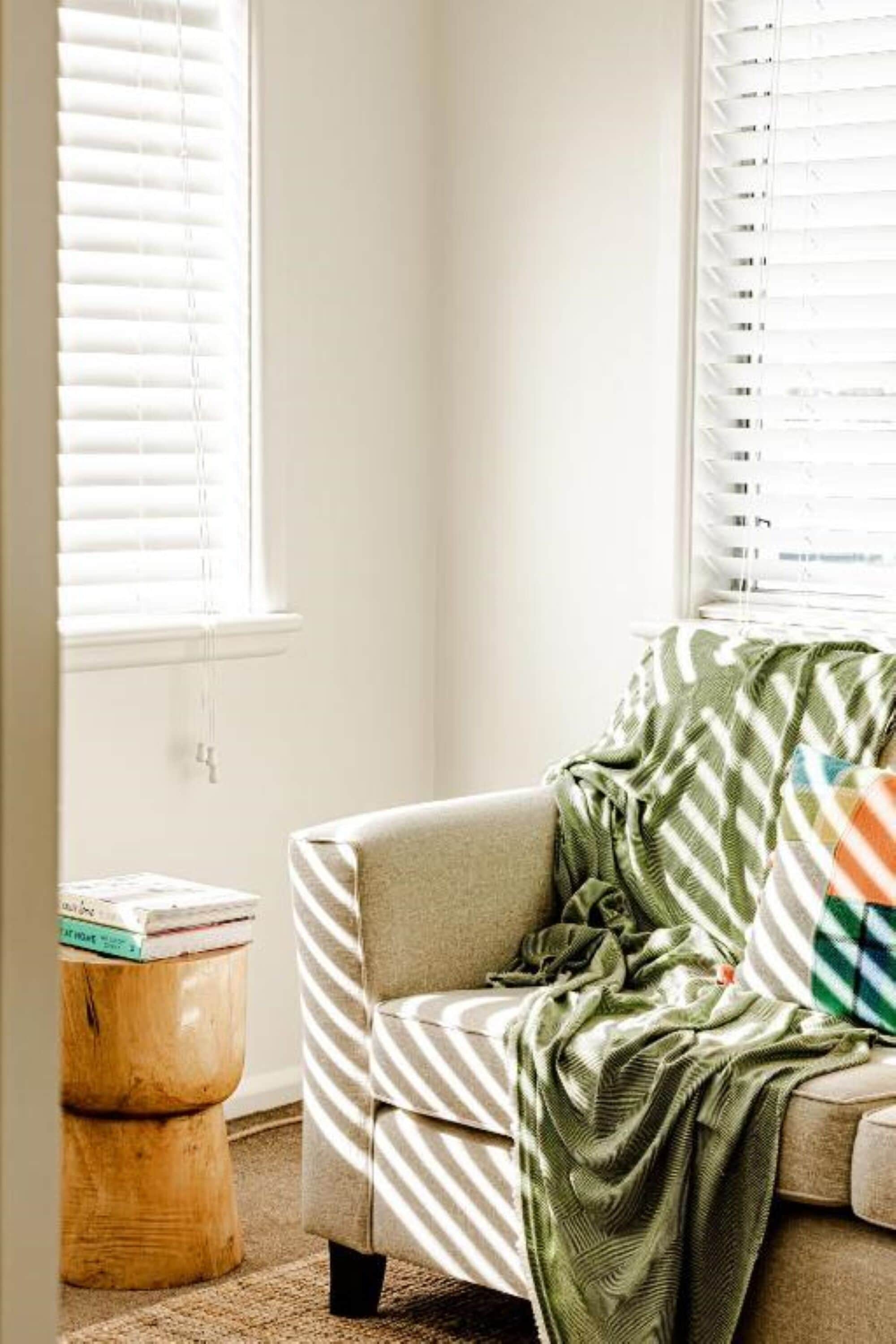 Eine Ecke eines Raumes mit einem beigen Sofa, einer grün-weiß gestreiften Decke und einem hölzernen Hocker mit Büchern.