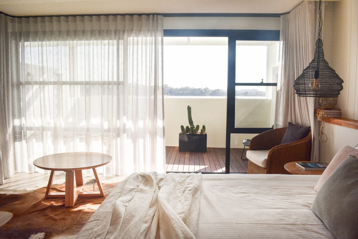 A bedroom with a bed, a round wooden table, a wicker chair, a potted cactus, and a view of the outdoors.