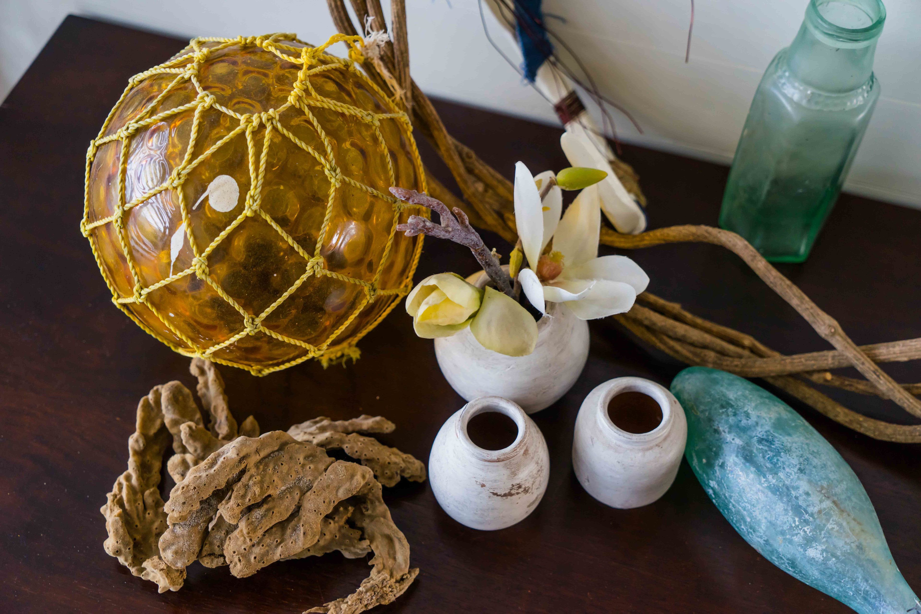 A decorative arrangement including a netted glass sphere, white ceramic vases, dried flowers, and a green bottle on a wooden surface.