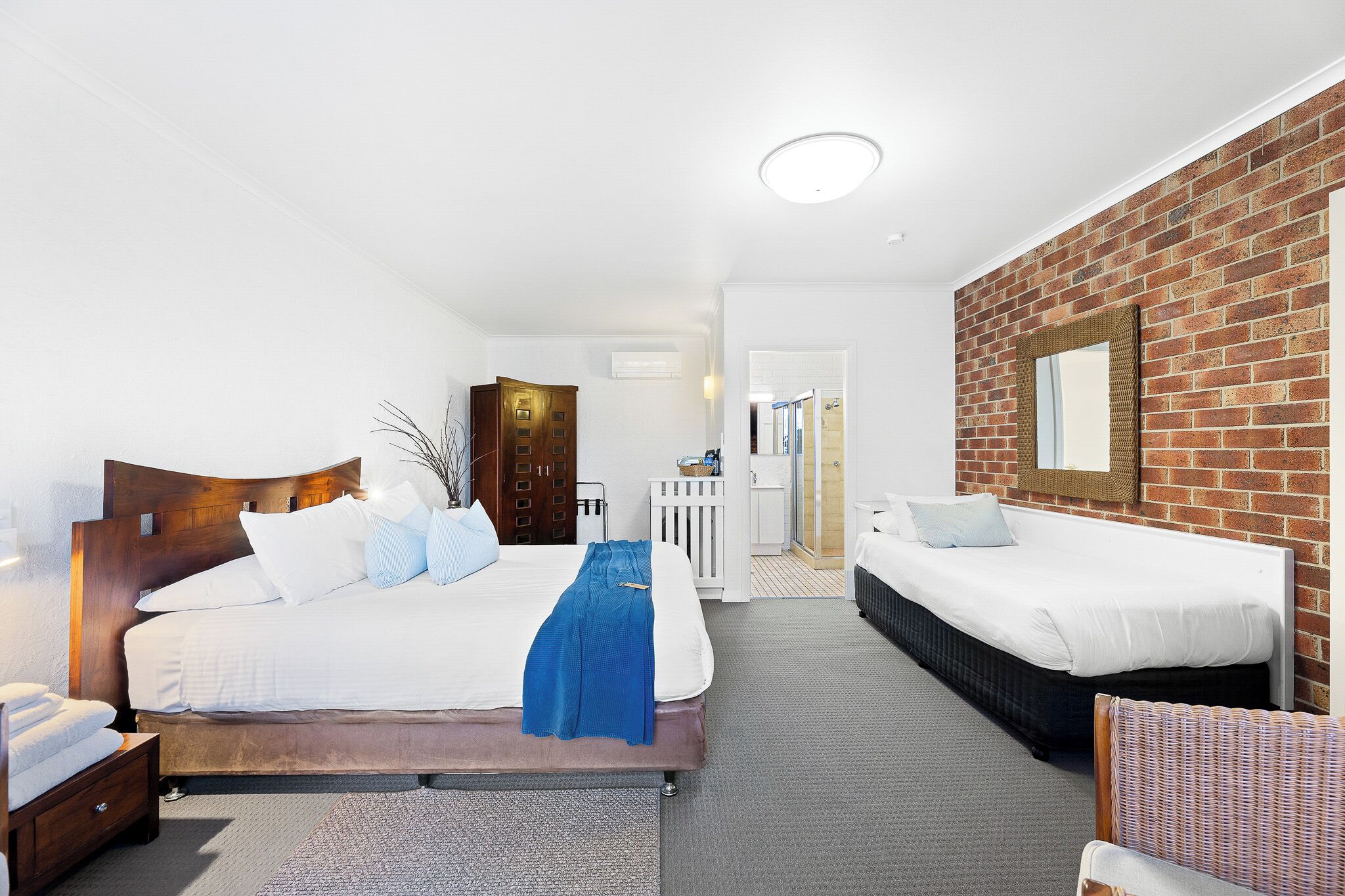 A hotel room with a large bed, a smaller bed, a wooden headboard, a white wall, a brick accent wall, a mirror, and a door leading to a bathroom.