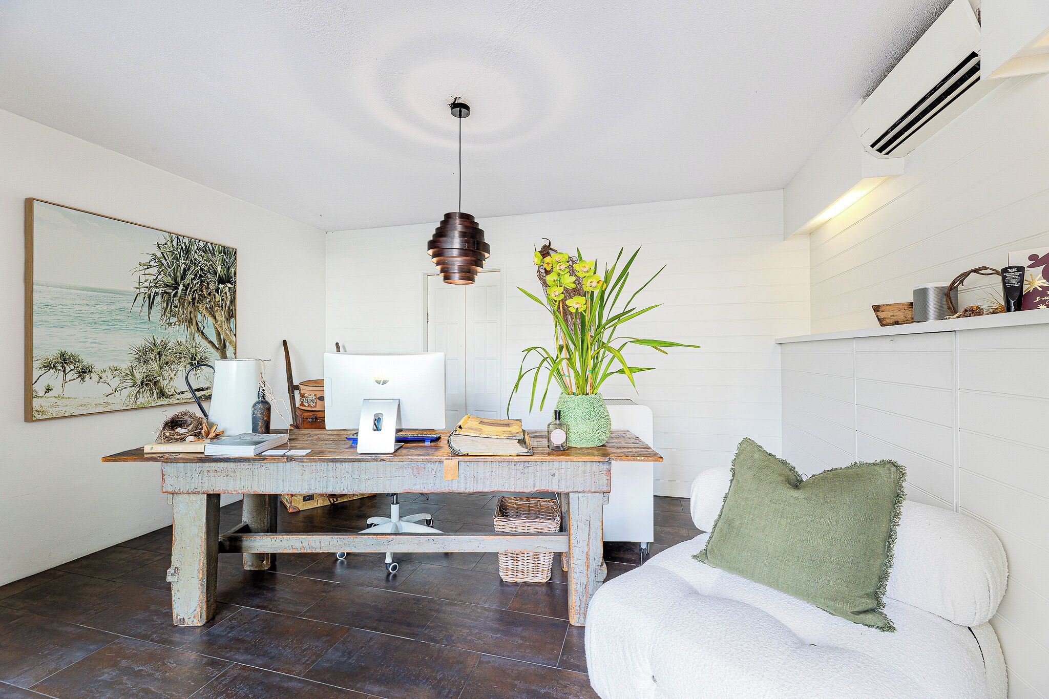 Una habitación con una mesa de madera, una lámpara colgante, un cuadro, una planta y un sofá blanco.