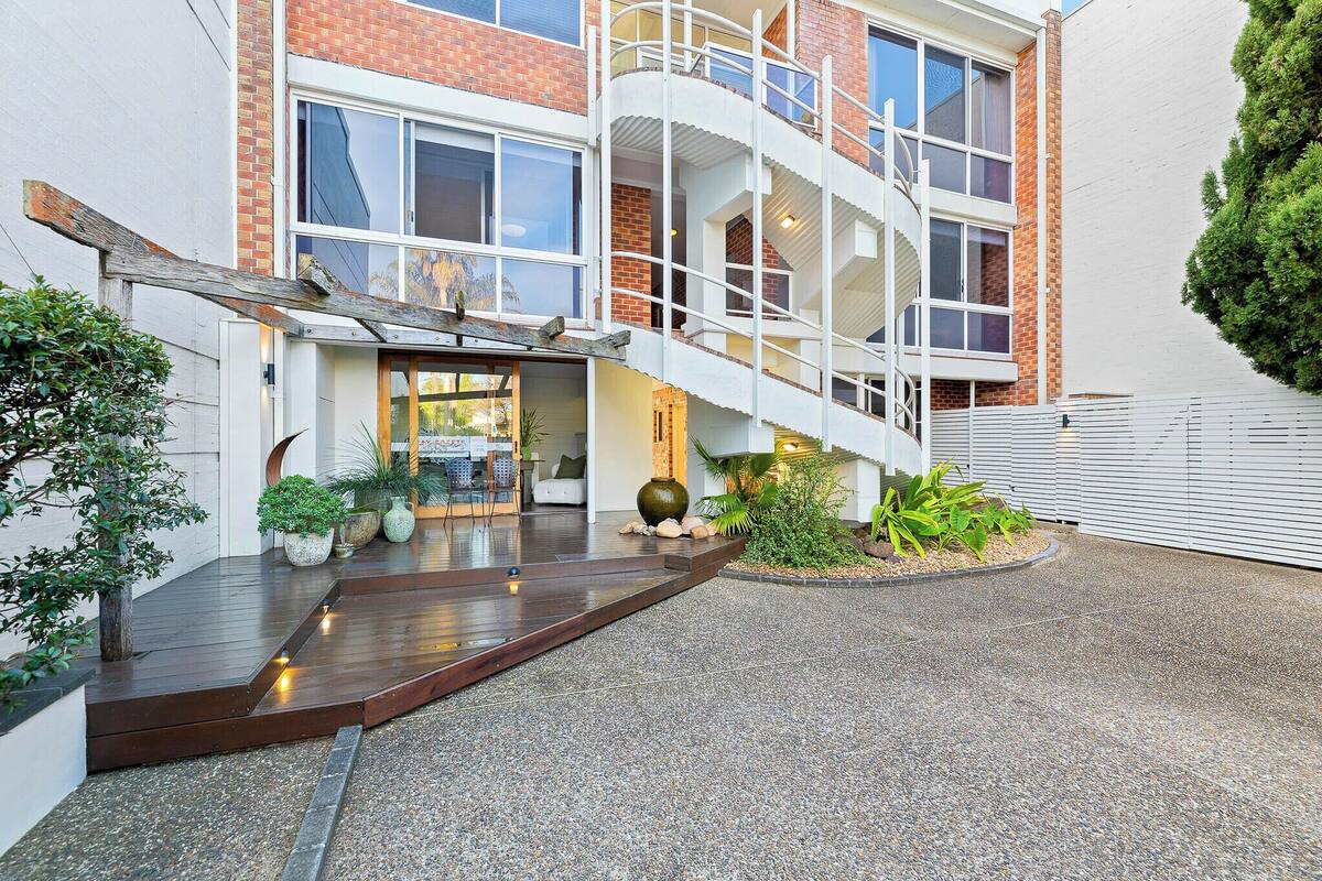 A multi-story building with a staircase leading to the entrance, surrounded by potted plants and a gravel driveway.