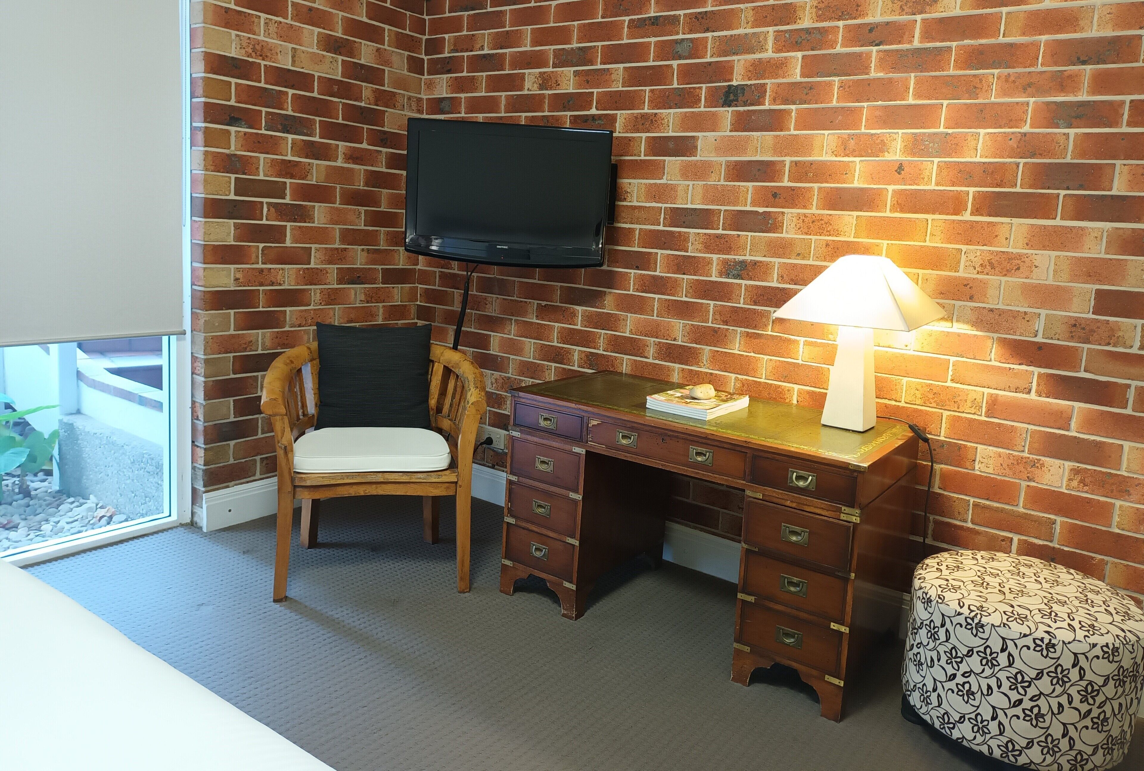 Una habitación con pared de ladrillo, un televisor sobre un escritorio de madera, una silla con cojín, una lámpara en el escritorio y un otoman estampado.