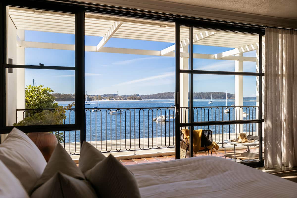 A hotel room with a large window overlooking a body of water, a balcony with a railing, and a bed with pillows.