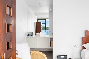A modern hotel room with a wooden door, a white bed, a white lamp, a wicker chair, and a view of the outdoors.
