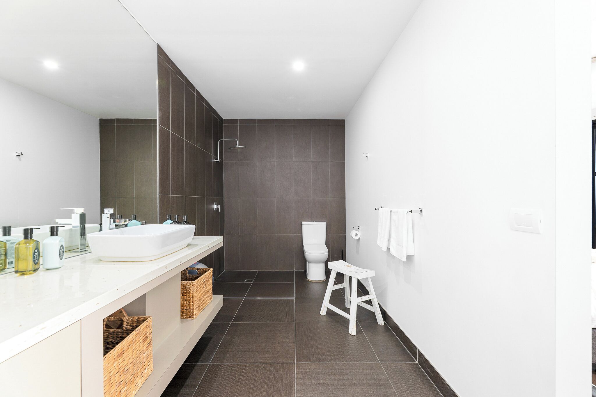 A modern bathroom with a large mirror, white sink, dark tiled shower, and a small stool.