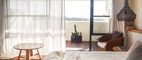Un dormitorio con una cama, una mesa redonda de madera, una silla de mimbre, un cactus en maceta y vistas al exterior.