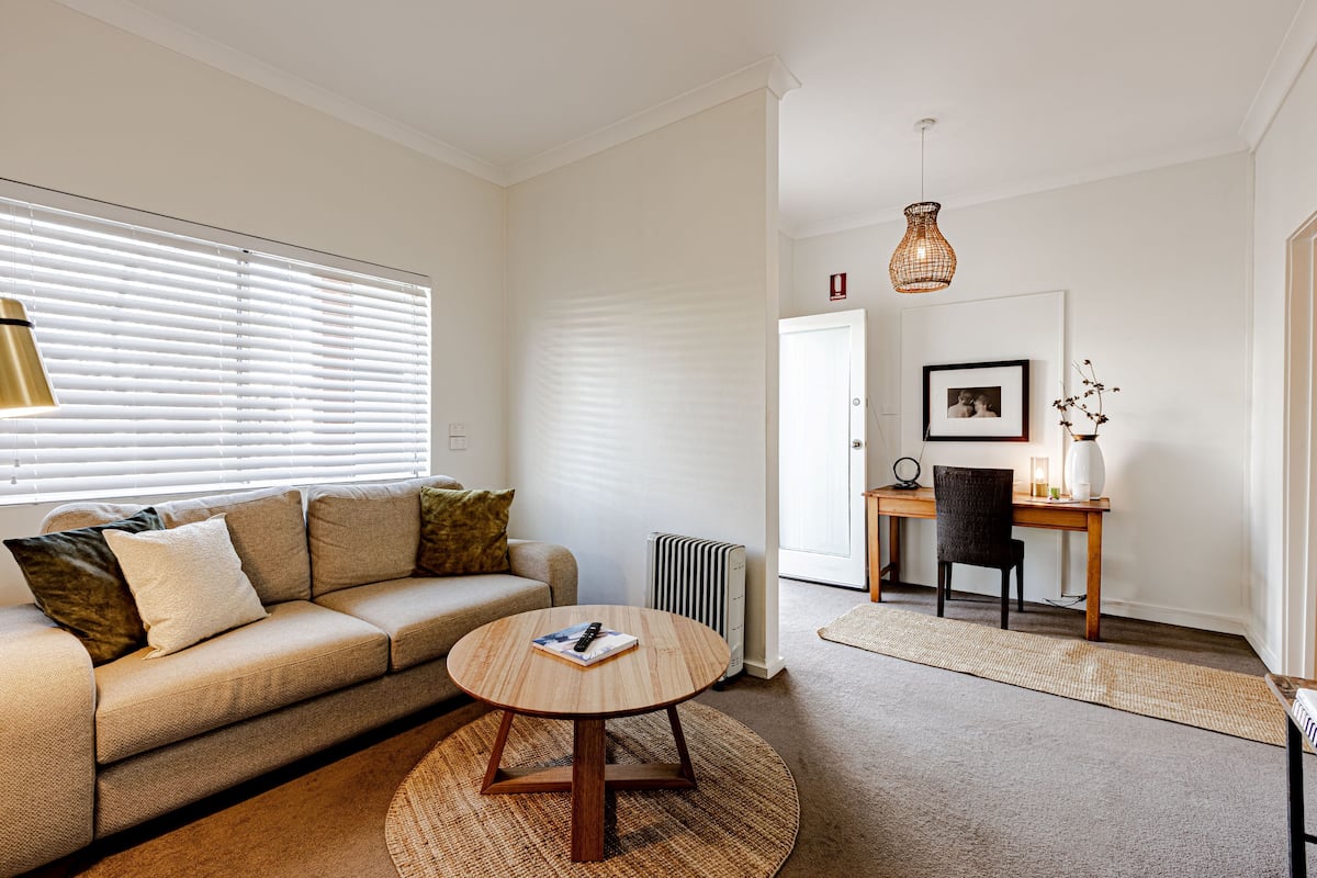 A living room with a sofa, a round coffee table, a radiator, a desk with a chair, and a hanging light fixture.