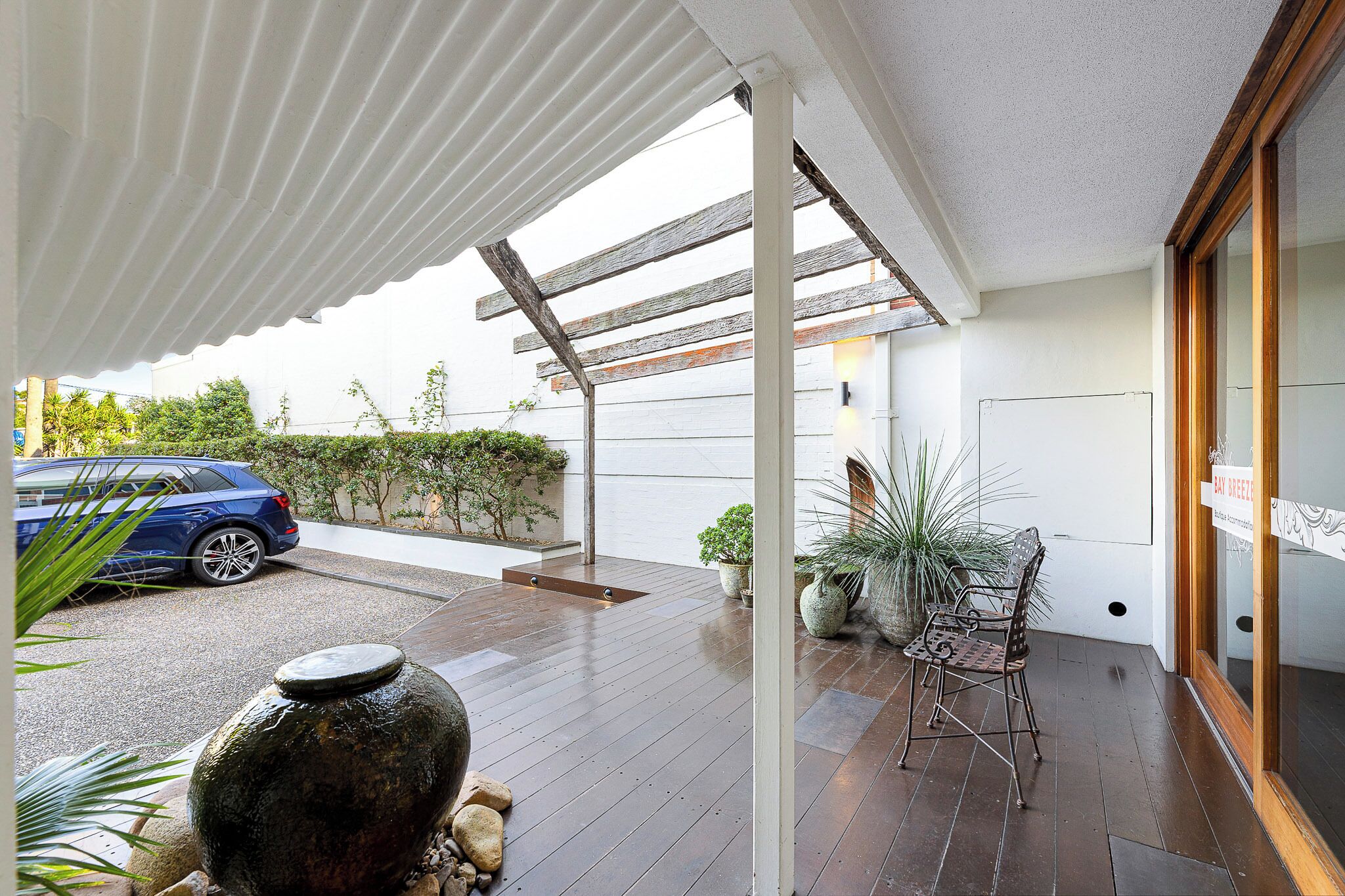 A covered outdoor area with a car parked nearby, wooden flooring, potted plants, and a chair.