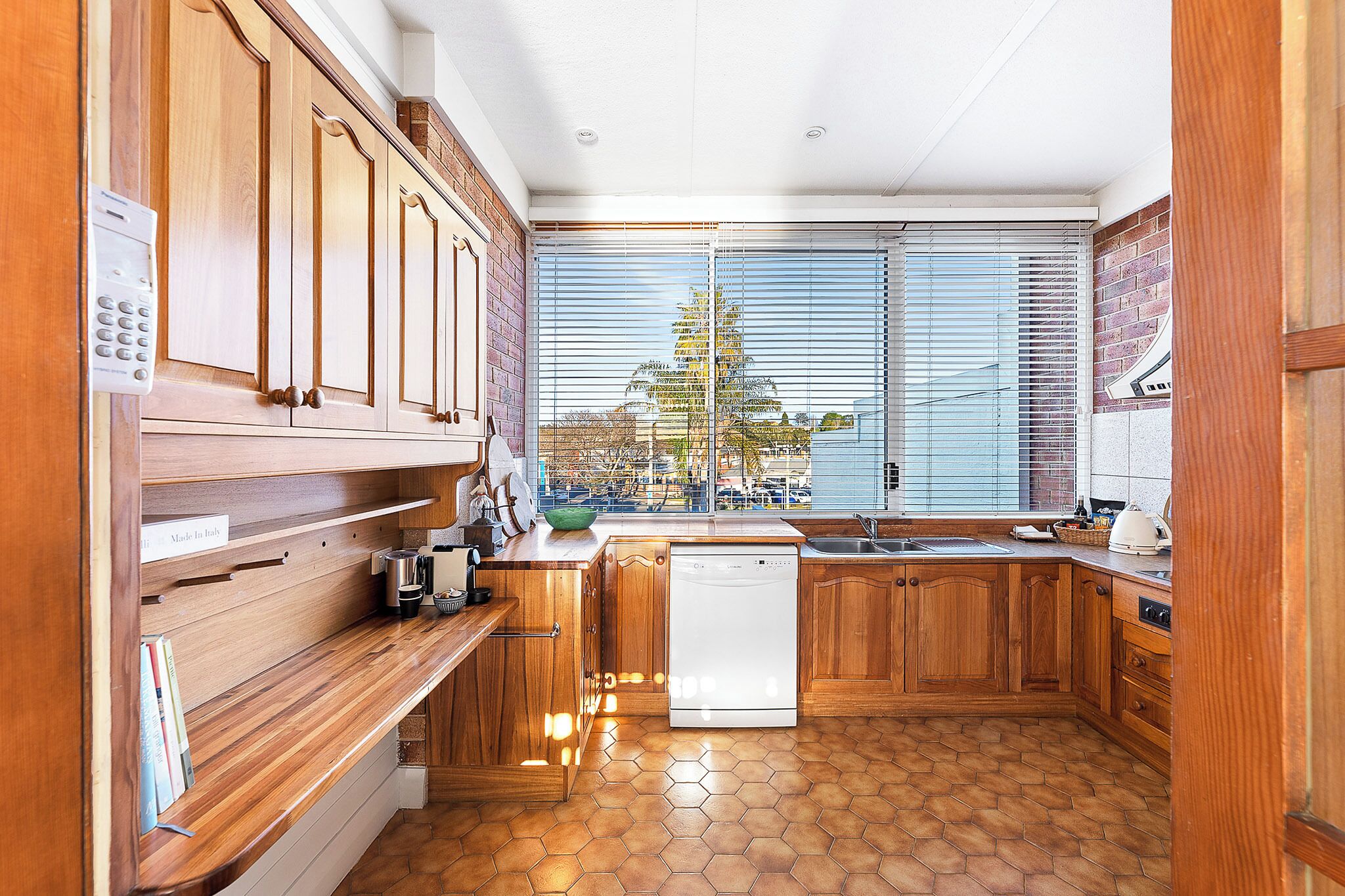 Une cuisine avec des meubles en bois, un lave-vaisselle, une fenêtre avec des stores et une vue sur les bâtiments environnants.