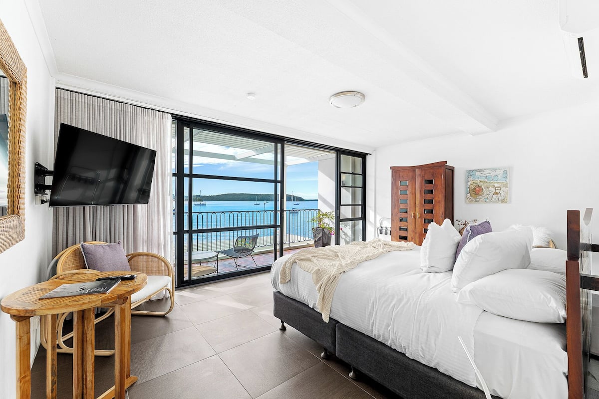 A bedroom with a bed, a TV, a chair, a table, and a view of the sea.