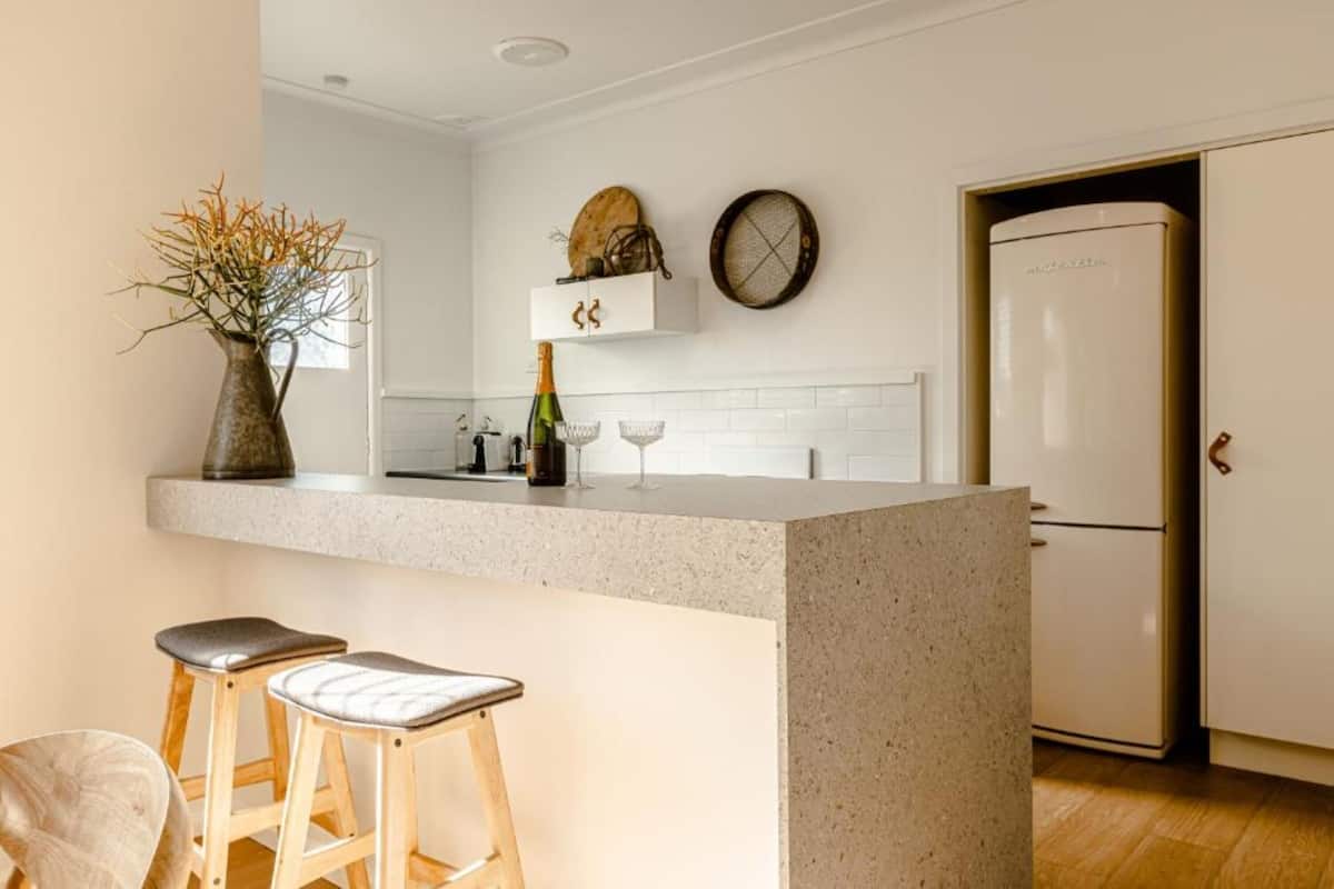 A kitchen with a central island, two stools, a vase with dried plants, a bottle of champagne, a wine glass, a clock, and a refrigerator.