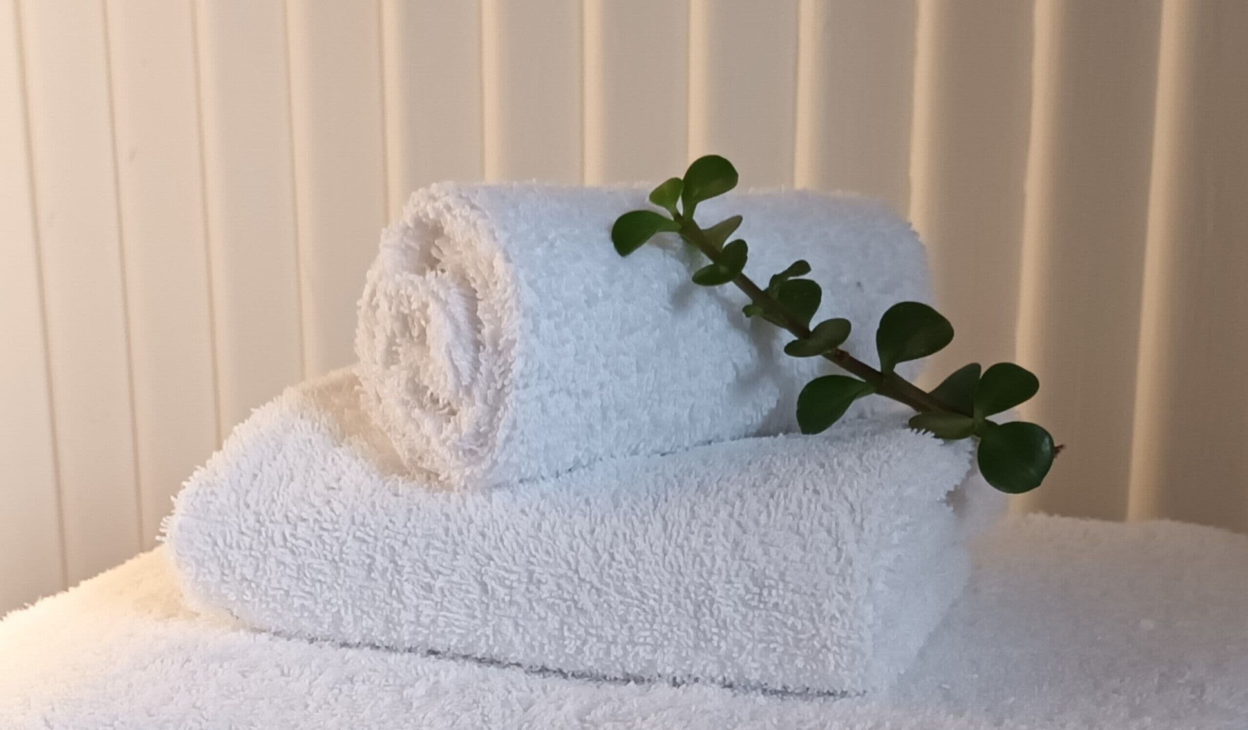 A neatly folded white towel with a green plant branch on top, placed on a white surface against a vertical striped background.