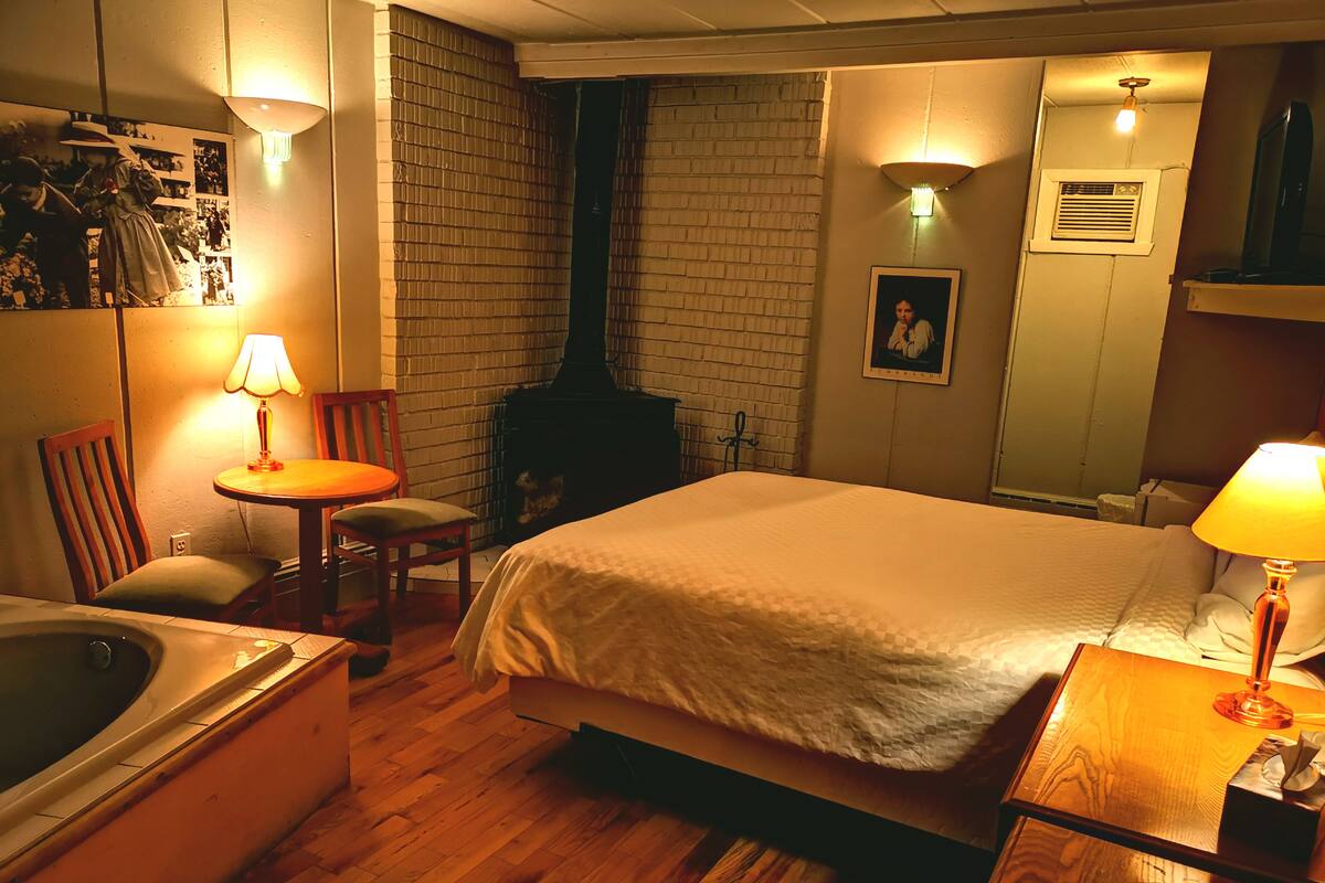 A cozy room with a bed, a small table with a lamp, a chair, a wooden floor, a brick wall, and a fireplace.