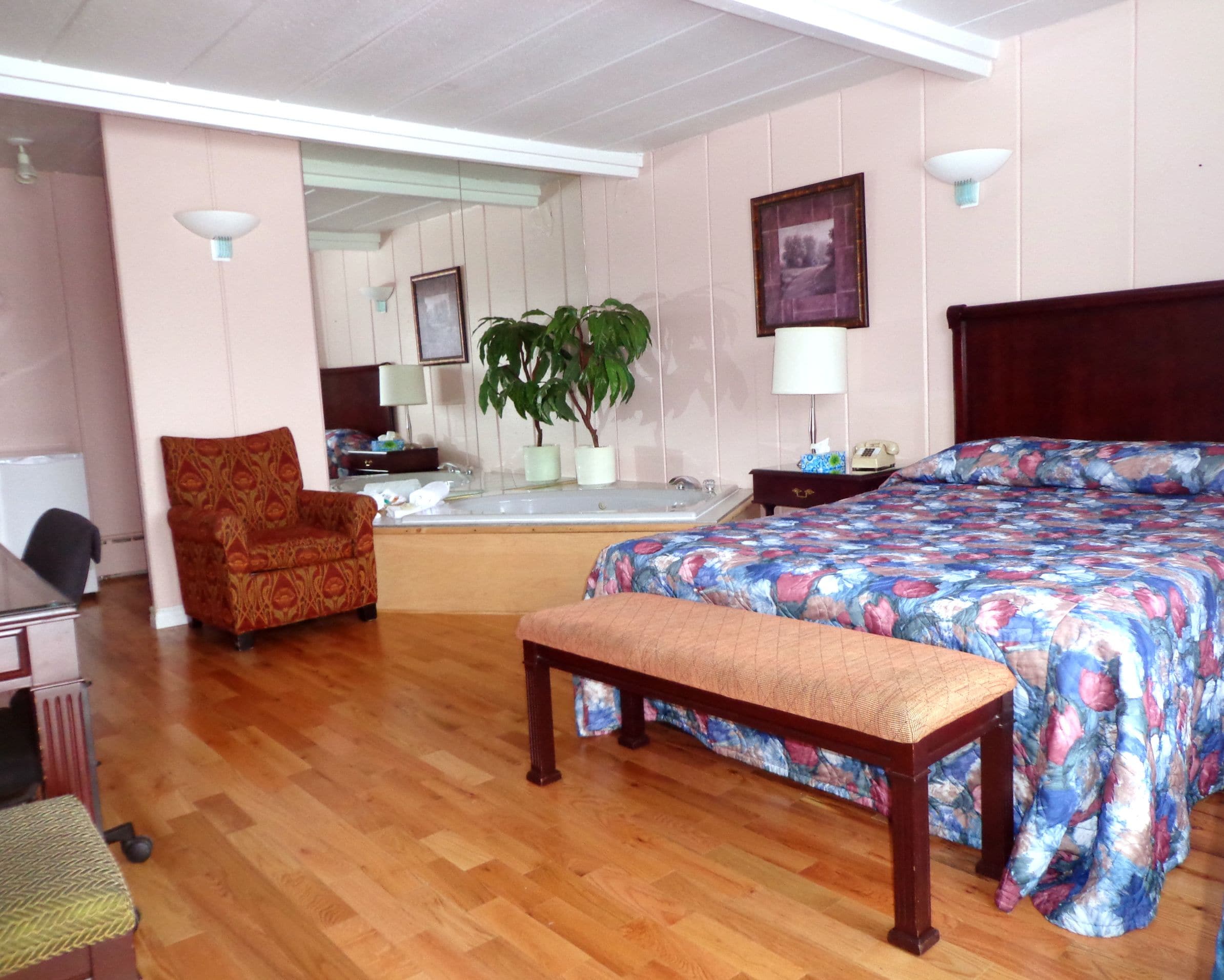 Une chambre d’hôtel comprenant un lit, un banc, une chaise, un bureau, un miroir, une plante et un coin salle de bain.