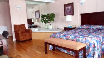 Une chambre d’hôtel comprenant un lit, un banc, une chaise, un bureau, un miroir, une plante et un coin salle de bain.